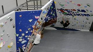 Image of students climbing on the NMT Bouldering Wall. The wall is made up of alternating blue and white panels.